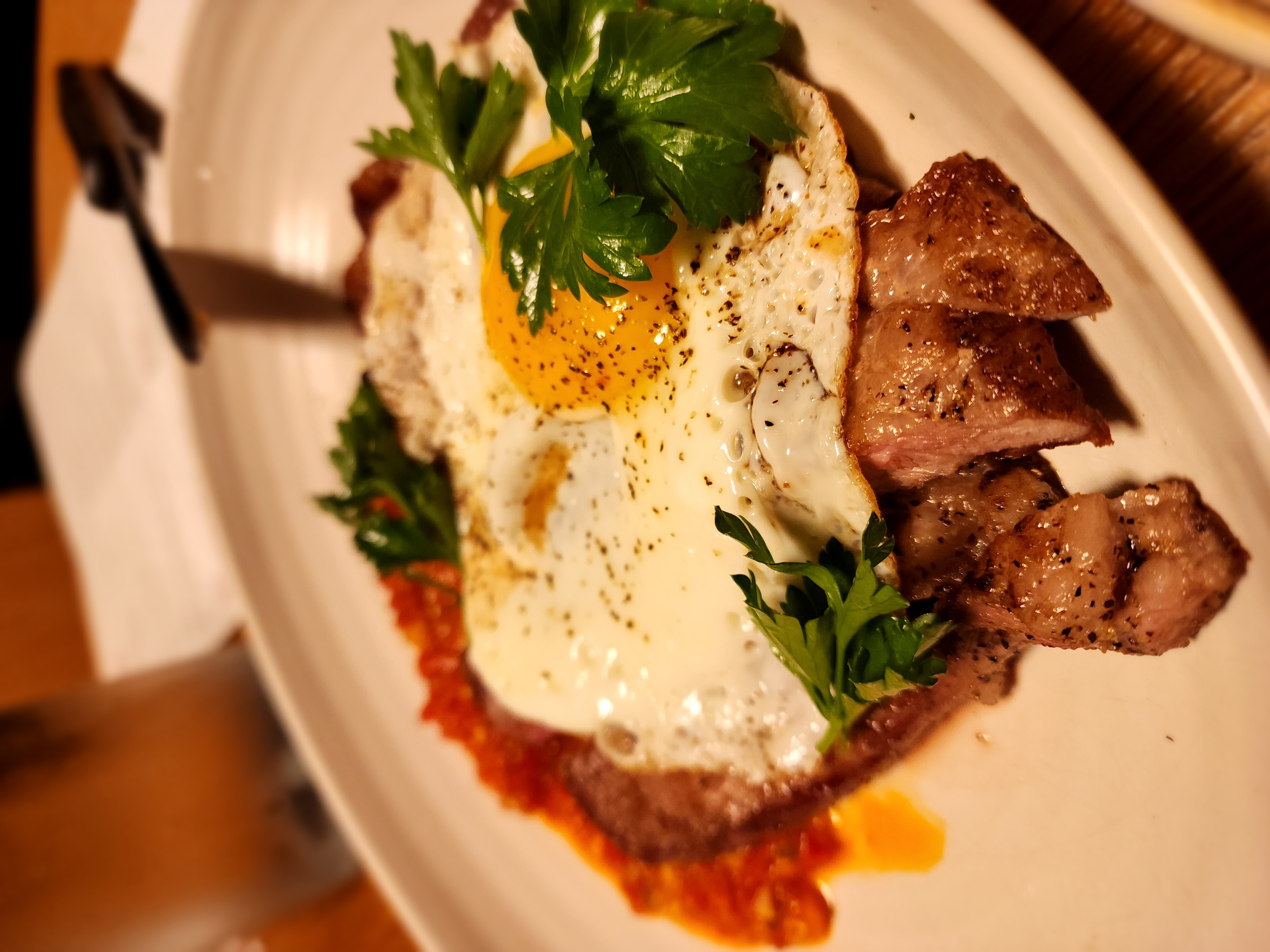 Slices of steak topped with a fried egg and fresh herbs on a white plate.