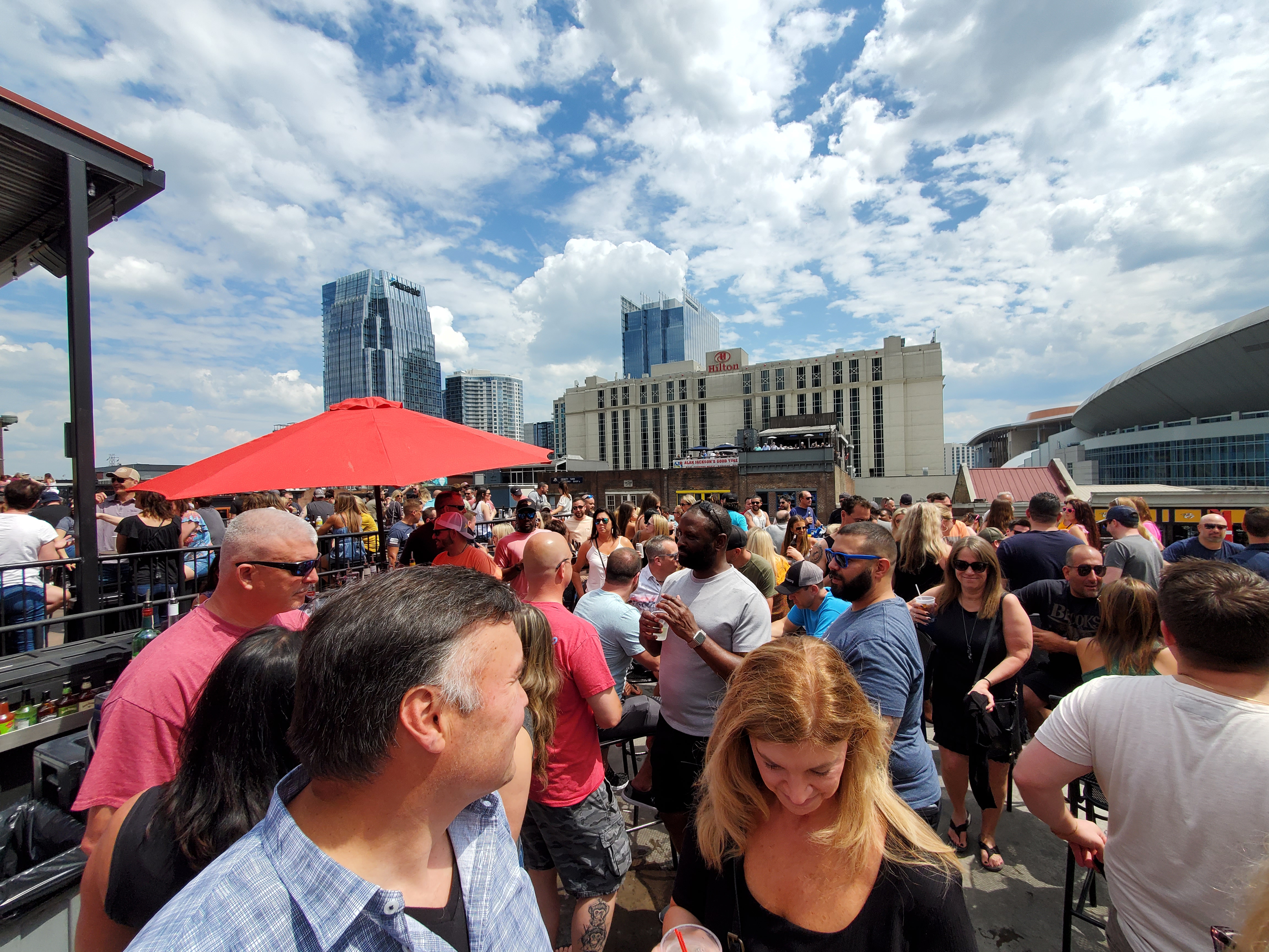 A daytime crowd gathers on a Broadway rooftop with the downtown skyline and Bridgestone Arena beyond.