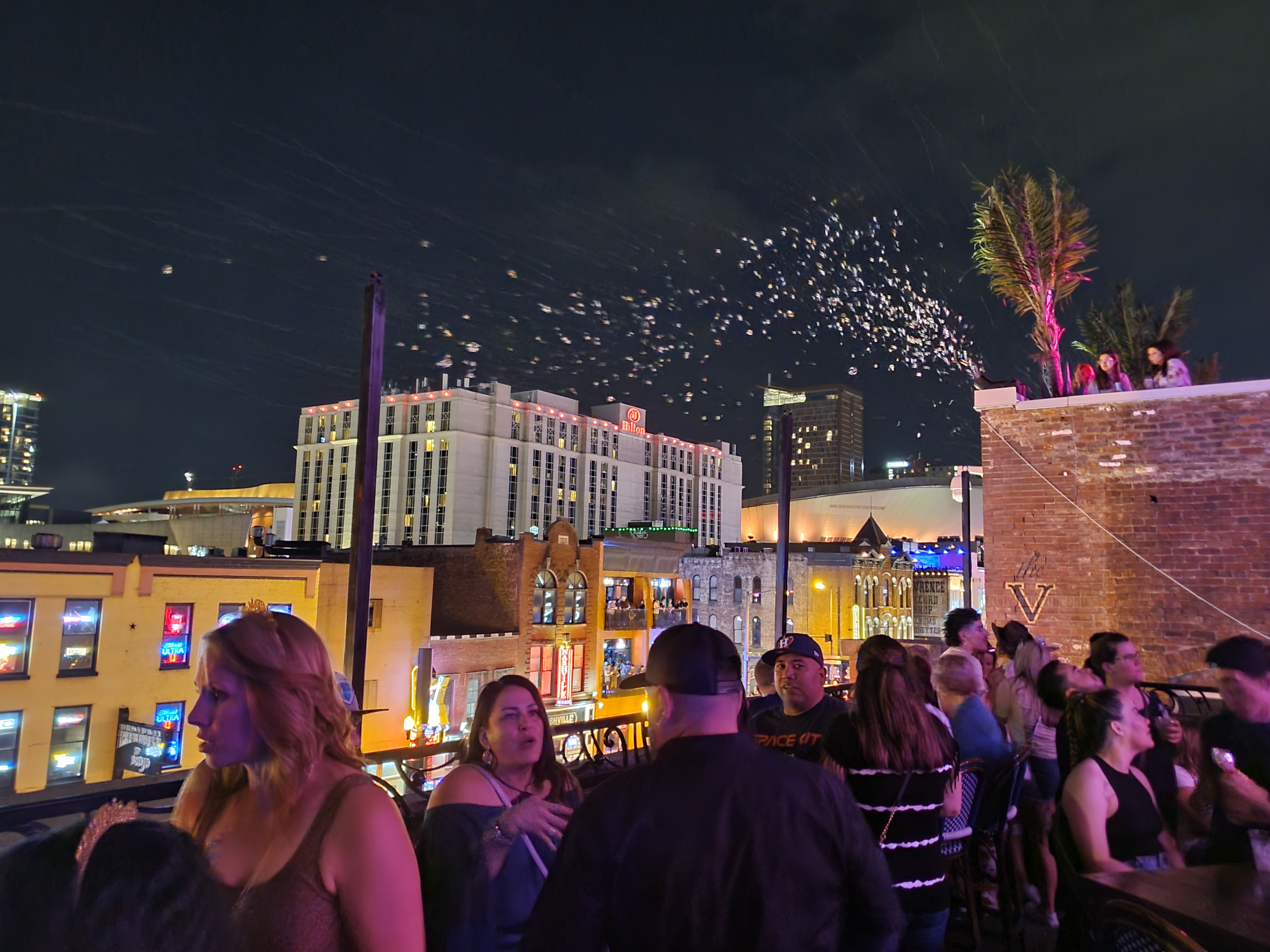 Visitors gather on a rooftop at night with illuminated Broadway buildings and neighboring terraces behind them.