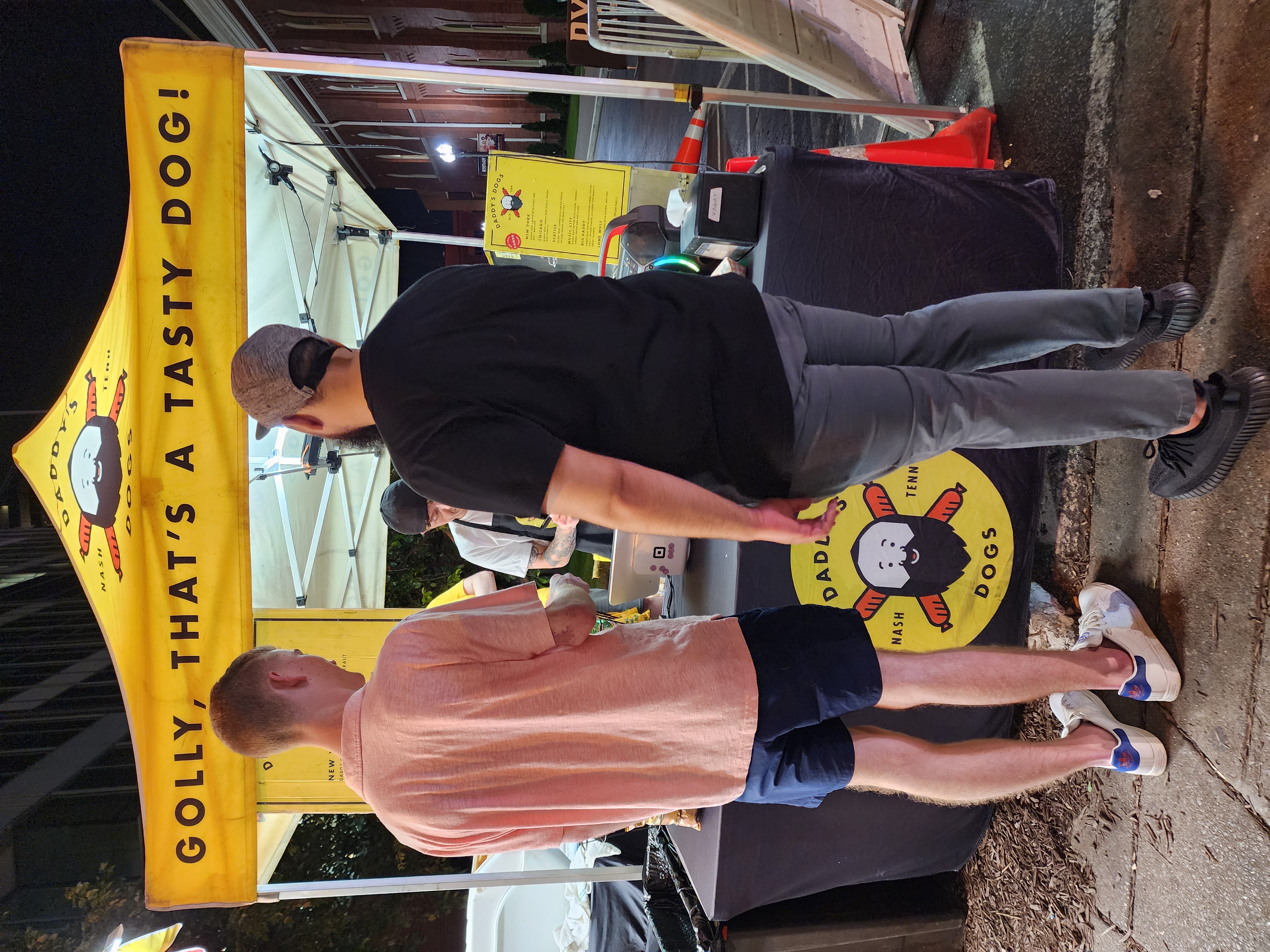 Customers order from a yellow Daddy's Dogs food stand near the Ryman at night.