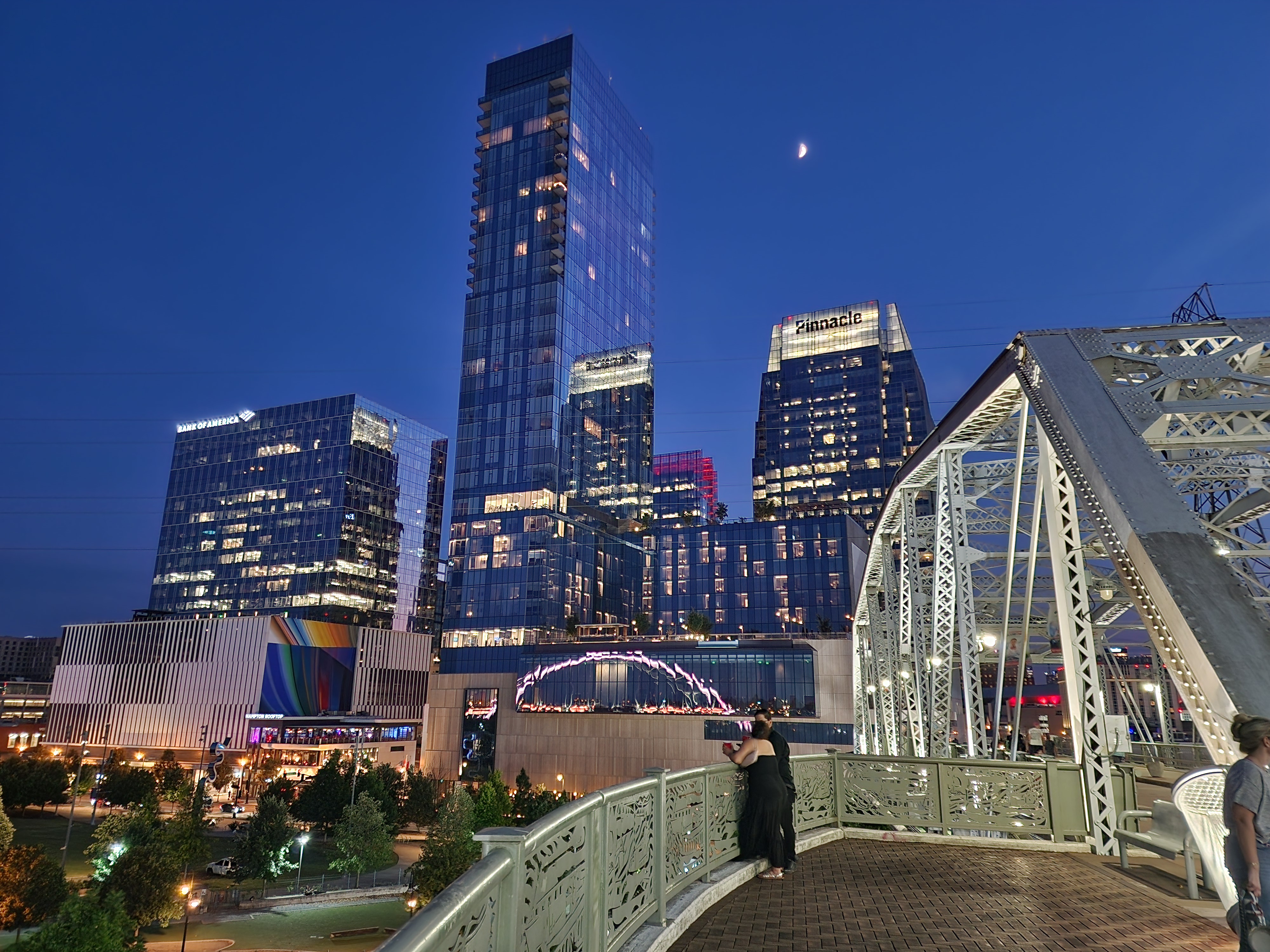 Pedestrians cross the John Seigenthaler Pedestrian Bridge beside the illuminated SoBro skyline at dusk.