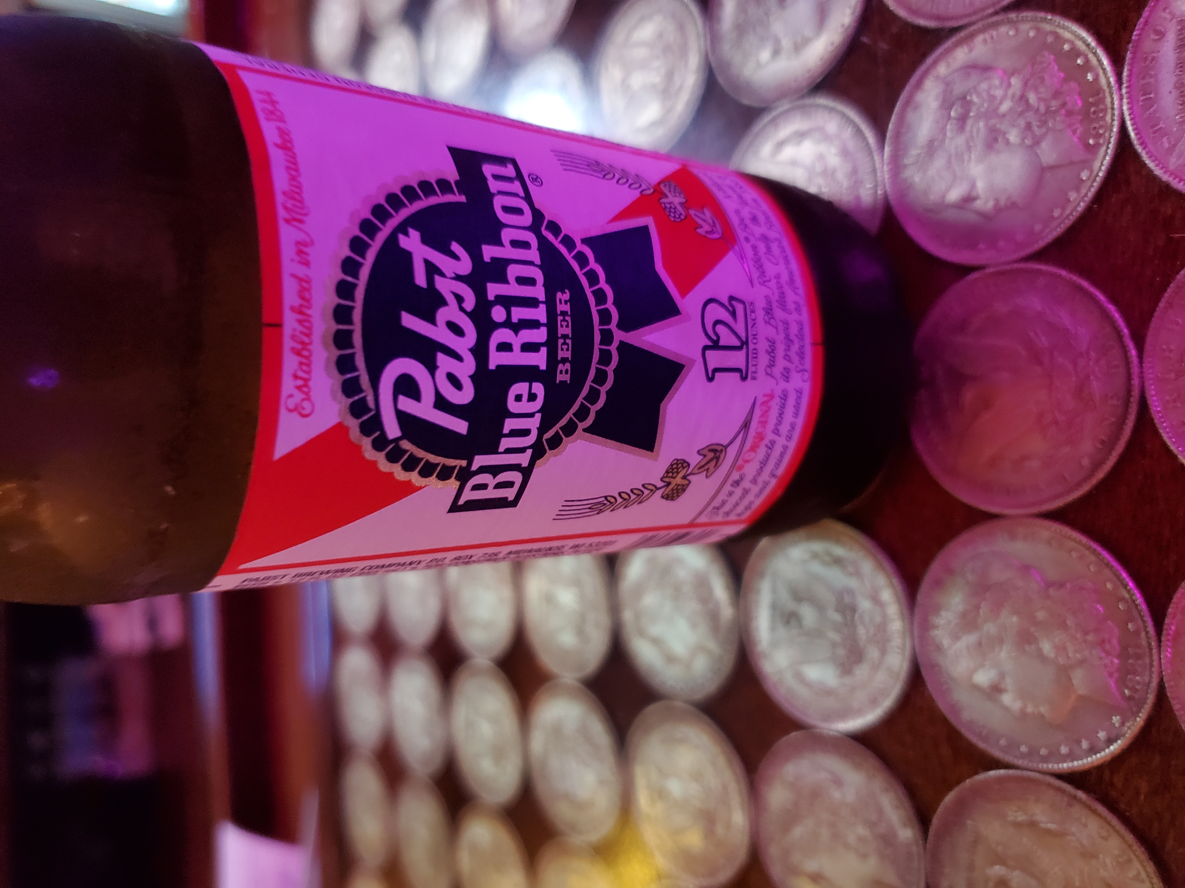 A Pabst Blue Ribbon bottle surrounded by coins on a bar.